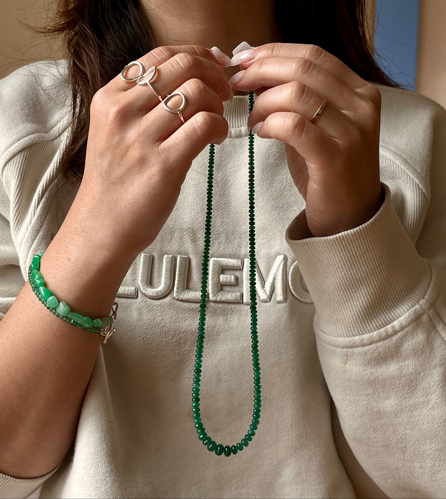 Person wearing a green beaded emerald necklace and jade bracelet, with a neutral background